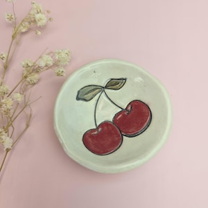 May include: A small, off-white ceramic dish with a hand-painted design of two red cherries with green leaves. The dish has a slightly irregular shape and sits on a pink surface. Decorative sprigs of white flowers are in the background.