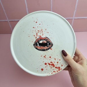 May include: A white ceramic plate featuring a vampire mouth design. The mouth is red and black with fangs. Red splatter-like paint is around the mouth. The plate is held by a hand with maroon nail polish.