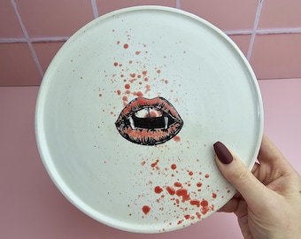 Könnte beinhalten: Ein weißer Keramikteller mit einem Vampir-Mund-Design. Der Mund ist rot und schwarz mit Fangzähnen. Rote, spritzartige Farbe befindet sich um den Mund herum. Der Teller wird von einer Hand mit weinrotem Nagellack gehalten.