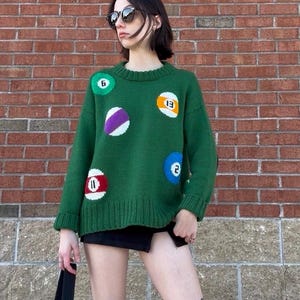 Könnte beinhalten: Ein grüner Strickpullover mit verschiedenen bunten Billardkugel-Designs. Der Pullover hat einen Rundhalsausschnitt und lange Ärmel. Die Billardkugeln sind in Grün-, Lila-, Rot- und Gelbtönen mit weißen Akzenten gehalten. Der Pullover wird mit einem schwarzen Rock getragen.
