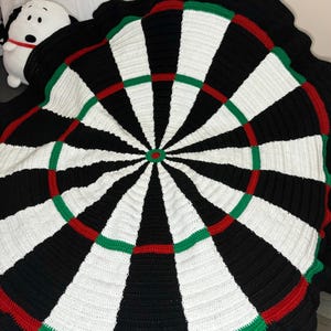 Op de afbeelding: Een gehaakt dartbord deken met zwart, wit, rood en groen garen. Het deken is rond en heeft een klassiek dartbord ontwerp.
