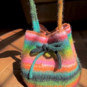Könnte beinhalten: Eine bunte gestreifte Häkeltasche mit schwarzen Kordeln. Die Tasche ist aus verschiedenen leuchtenden Farben gefertigt, darunter Pink, Orange, Grün und Blau.