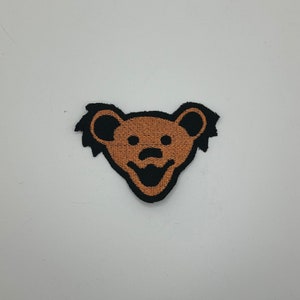 May include: A brown and black embroidered patch of a smiling bear with a black outline. The bear has a round nose and large, round ears.
