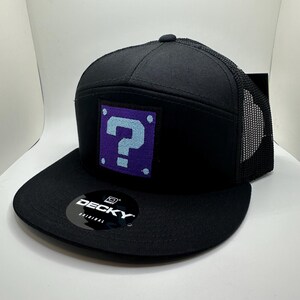 May include: Black trucker hat with a purple and blue question mark block on the front. The hat has a black mesh back and a black brim. The hat has a "DECKY ORIGINAL" label on the brim.