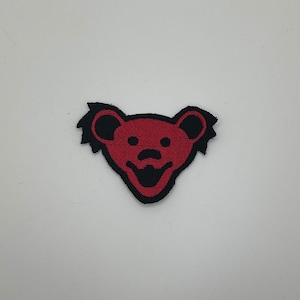 May include: Red and black embroidered patch of a smiling bear with a fuzzy outline.