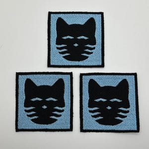 May include: Three black cat face iron-on patches on a white background. The cat faces are outlined in black and have white whiskers. The patches are on a light blue background.