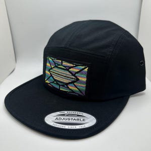May include: Black 5-panel hat with a colorful geometric design embroidered on the front. The hat has an adjustable strap with the text "The Authentic Yupoong Adjustable Yupoong Since 1974".