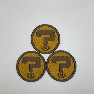 May include: Three circular patches with a brown question mark and eye design on a gold background. The patches have a brown border and are arranged in a triangular pattern against a white backdrop. The patches are likely for clothing or accessories.