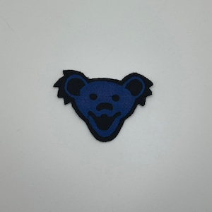 May include: A blue and black embroidered patch featuring a bear's face with a smiling mouth.