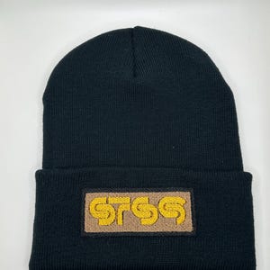May include: Black beanie with a brown rectangular patch featuring the embroidered text "STSS" in yellow. The beanie is made of a knitted material and has a folded brim.