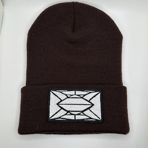Puede incluir: Un gorro de punto marrón con un diseño geométrico blanco bordado en la parte delantera.