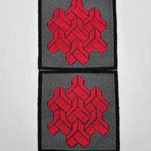 May include: Two square patches with a gray background and a black border. Each patch features a red, interwoven geometric design. The patches are likely for clothing or accessories, offering a unique, textured aesthetic.