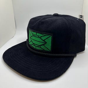 May include: A black corduroy baseball cap with a green geometric design patch. The cap has a black rope trim around the brim.