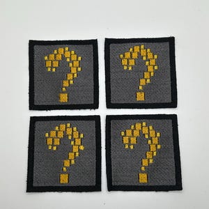 May include: Four square patches with a gray background and a black border. Each patch features a yellow pixelated question mark design. The patches are likely embroidered or sewn.
