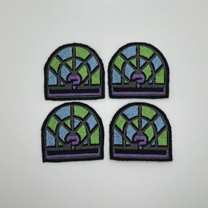 May include: Four embroidered patches with a stained-glass window design. Each patch features a black border, with sections of blue, green, and gray, and a purple central element. The patches are arranged in a square formation.