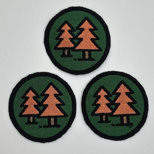 Op de afbeelding: Drie ronde patches met een groene achtergrond en een zwarte rand. Elke patch heeft twee oranje dennenbomen. Deze patches zijn geschikt voor kleding of accessoires en zijn ideaal voor outdoorliefhebbers.