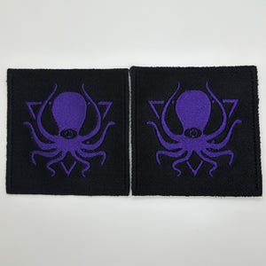 May include: Two black fabric patches with purple embroidered octopus designs inside a triangle. The octopus has one eye in the center of its head.
