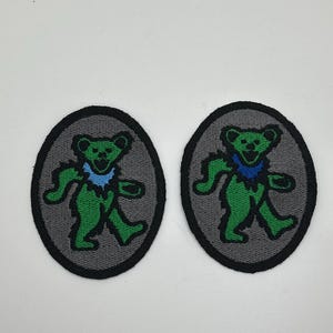 May include: Two embroidered patches featuring a green bear with a blue collar, set against a gray oval background. The bear is in a walking pose, with a black border around the oval. These patches are suitable for clothing or accessories.
