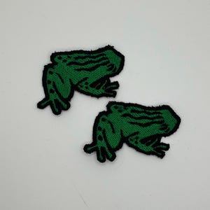 Op de afbeelding: Twee groene kikkerpatches met zwarte contouren. De patches zijn geborduurd met een gedetailleerd kikkerontwerp, met een getextureerde huid en gedefinieerde ledematen. De kikkers zijn in dezelfde richting geplaatst, voor kleding of accessoires.