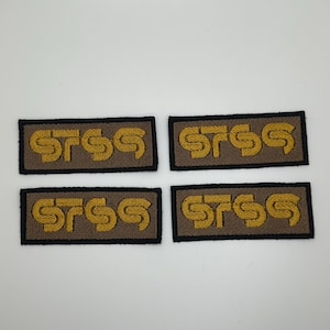 May include: Four rectangular patches with the letters "STSS" embroidered in gold thread on a brown background. Each patch has a black border. The patches are arranged in two rows of two.