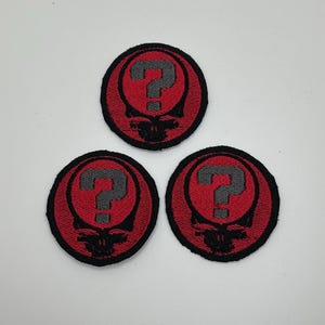 May include: Three circular patches with a red background and a black border. Each patch features a gray question mark over a stylized black skull design. The patches are likely for clothing or accessories.