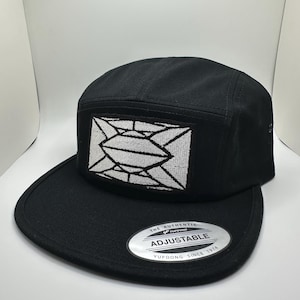 May include: Black 5-panel cap with a white geometric design patch on the front. The cap has an adjustable strap with a silver label that reads "The Authentic Yupoong Adjustable Yupoong Since 1974".