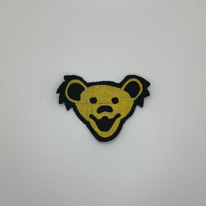 May include: A yellow and black embroidered patch featuring a smiling bear with a black nose and eyes. The bear has a black outline and black fur around its head.