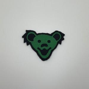 May include: Green and black embroidered patch of a bear with a smiling face. The bear has a black outline and a green face.