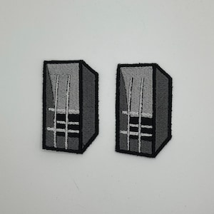 May include: Two embroidered patches with a geometric design. Each patch is a dark grey, three-dimensional shape with a lighter grey interior and white vertical lines. The patches are outlined in black.