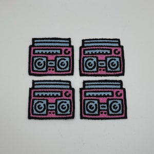 May include: Four embroidered patches featuring a retro boombox design. Each patch has a pink body with blue accents and black outlines. The boomboxes have speaker details and antenna.