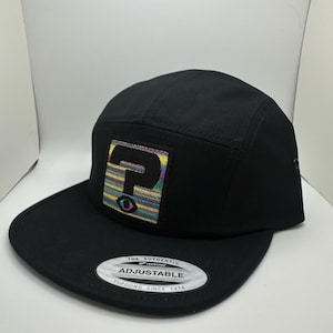 May include: Black 5-panel hat with a colorful question mark patch and an adjustable strap with the text "The Authentic Yupoong Adjustable Since 1974".