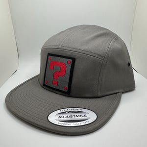 May include: Gray 5-panel cap with a red question mark patch and an adjustable strap. The strap has the text "The Authentic Yupoong Adjustable Yupoong Since 1974".