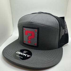 Puede incluir: Una gorra de camionero gris y negra con un signo de interrogación rojo en un cuadrado negro en la parte delantera. La gorra tiene una parte trasera de malla negra y una etiqueta negra "DECKY ORIGINAL" en el lateral.