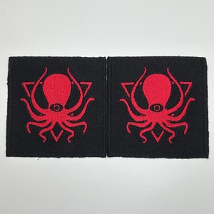 May include: Two black fabric patches with red embroidered octopus designs inside a triangle. The octopus has one eye.