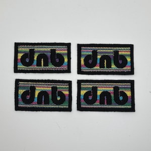 Op de afbeelding: Vier rechthoekige patches met de letters "dnb" geborduurd in zwart op een kleurrijke gestreepte achtergrond. De patches hebben een zwarte rand en zijn gerangschikt in een raster van twee bij twee.