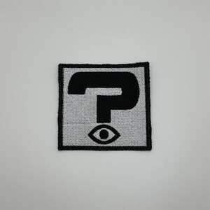 May include: A black and white embroidered patch with a question mark and an eye. The patch is rectangular and has a white background with black stitching.
