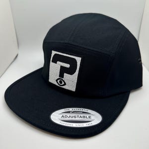May include: Black 5-panel baseball cap with a white eye logo and the text "THE AUTHENTIC YUPOONG ADJUSTABLE YUPOONG SINCE 1974" on the back.