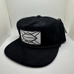 May include: A black corduroy baseball cap with a white geometric design embroidered on the front. The cap has a black brim and a black rope detail around the crown.