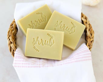May include: Three green bars of soap with the word "shrub" embossed on each bar. The soap bars are in a woven basket lined with a white and pink striped towel.