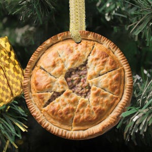 May include: A round, baked pie ornament with a golden crust and filling, cut into wedges. The ornament is suspended by a gold ribbon against a backdrop of a Christmas tree. The pie's filling is visible in the center.