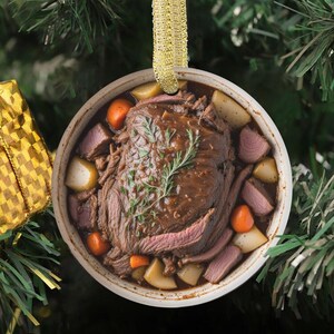 May include: A circular Christmas decoration depicting a pot roast meal. The roast is surrounded by potatoes, carrots, and gravy. A sprig of rosemary sits on top. The ornament hangs from a gold ribbon.