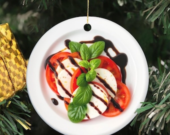 May include: A round, white ceramic ornament featuring a caprese salad. The salad includes sliced red tomatoes, mozzarella cheese, fresh basil, and balsamic glaze. A gold string is attached for hanging. The ornament is set against a backdrop of Christmas tree branches.