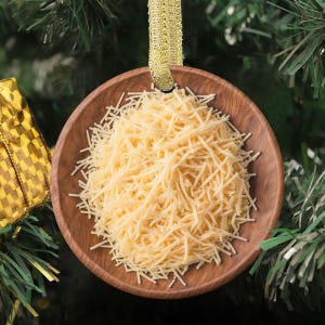May include: A wooden Christmas ornament shaped like a bowl filled with shredded pasta. The ornament hangs from a gold ribbon against a backdrop of a Christmas tree. The pasta is a light yellow color.