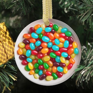 Op de afbeelding: Een rond, wit keramisch ornament met een levendige, hoge resolutie print van een grote hoop kleurrijke jelly beans in tinten rood, oranje, geel, groen, blauw en paars. Het ornament hangt aan een glinsterende gouden lintlus.