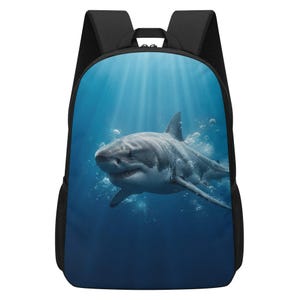 May include: A black backpack featuring a large, high-resolution graphic print of a great white shark swimming underwater. The shark is surrounded by bubbles with light rays filtering down through the deep blue ocean water from the surface above.