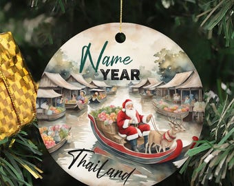 May include: Round Christmas ornament featuring Santa Claus in a boat with a reindeer, navigating a Thai floating market. The ornament has the words "Name", "Year", and "Thailand" on it. The ornament is hanging from a gold string.