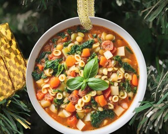 May include: A round, white ceramic ornament featuring a detailed image of minestrone soup with pasta, carrots, beans, leafy greens, and a fresh basil garnish in the center, suspended by a shimmering gold ribbon against a green pine background.