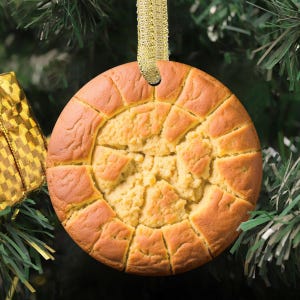 May include: A round, golden-brown cookie-shaped Christmas ornament with a textured, cracked surface pattern. The ornament features a light tan center and a darker, segmented outer ring, suspended by a shimmering gold ribbon loop.
