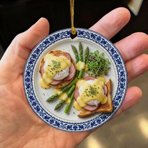 以下が含まれることがあります： ハム、オランデーズソース、新鮮なアスパラガス、スプラウトを添えたイングリッシュマフィンのエッグベネディクト2人前が描かれた、青と白のフローラルボーダーが特徴の円形セラミックオーナメント。