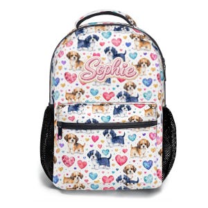 May include: White backpack with a pattern of puppies, hearts, and colorful dots. The name "Sophie" is written in pink script across the front. The backpack has black mesh side pockets and a top handle.